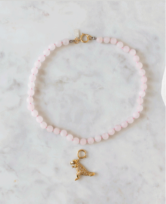 A pink beaded necklace featuring a gold dinosaur charm from WALD Berlin’s “DIY Schmuck-Workshop x MINI im KaDeWe Berlin” is showcased on a light marble surface, perfect for jewelry-making enthusiasts.