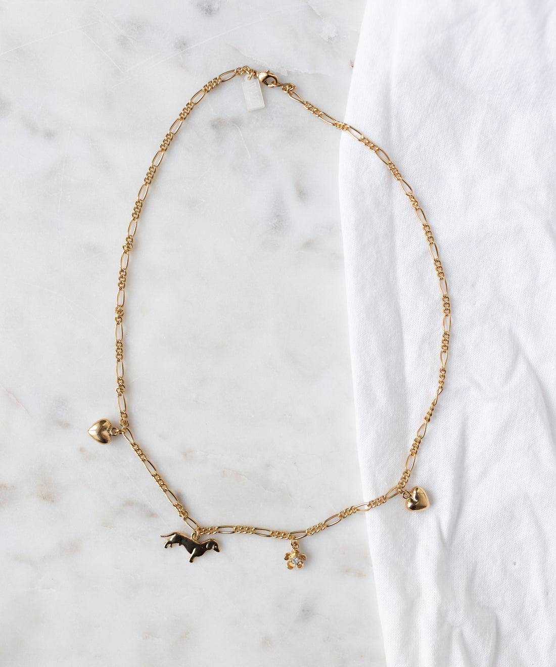 The WALD Berlin SAMPLE SALE Charm Necklace Z Edition, an 18k gold-plated chain with heart, horse, and flower charms, is shown on a white marble surface beside a white cloth.