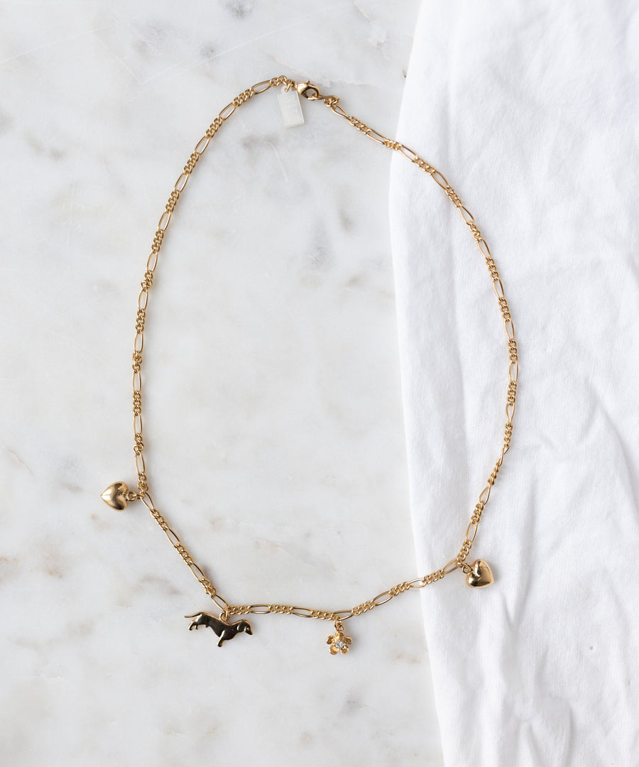 The WALD Berlin SAMPLE SALE Charm Necklace Z Edition, an 18k gold-plated chain with heart, horse, and flower charms, is shown on a white marble surface beside a white cloth.
