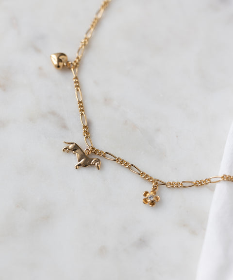 The WALD Berlin SAMPLE SALE Charm Necklace Z Edition features 18k gold plating, a heart charm, a small dog-shaped charm, and a flower charm with a clear stone, shown on white marble.