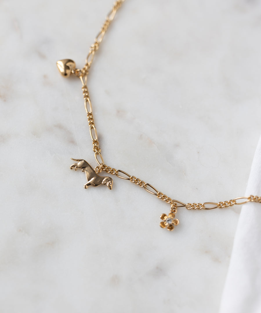 The WALD Berlin SAMPLE SALE Charm Necklace Z Edition features 18k gold plating, a heart charm, a small dog-shaped charm, and a flower charm with a clear stone, shown on white marble.