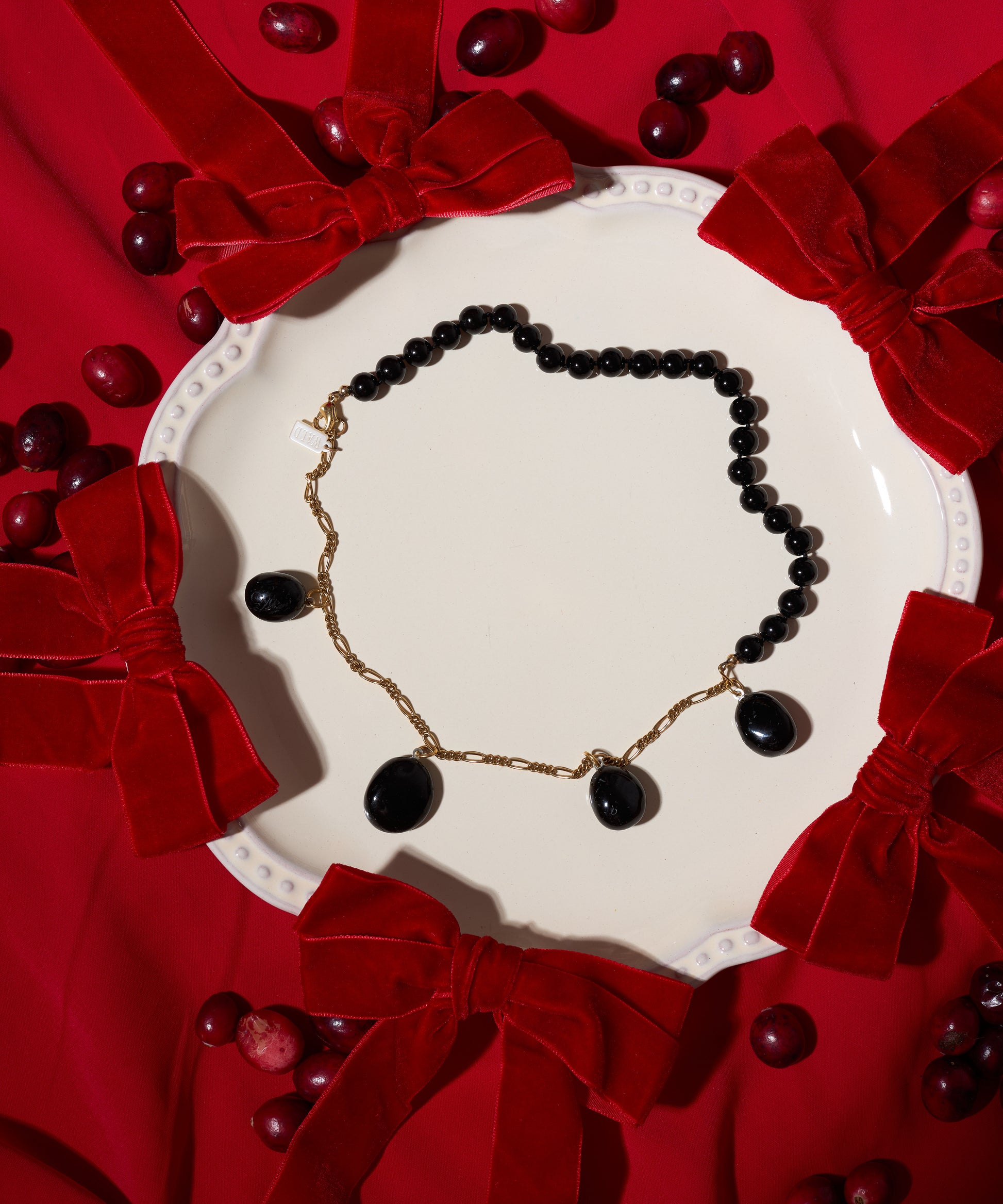 A white plate displays the WALD Berlin Collection Noir Pas De Deux Black Drop Necklace, surrounded by red velvet bows and scattered cranberries on a red fabric background.