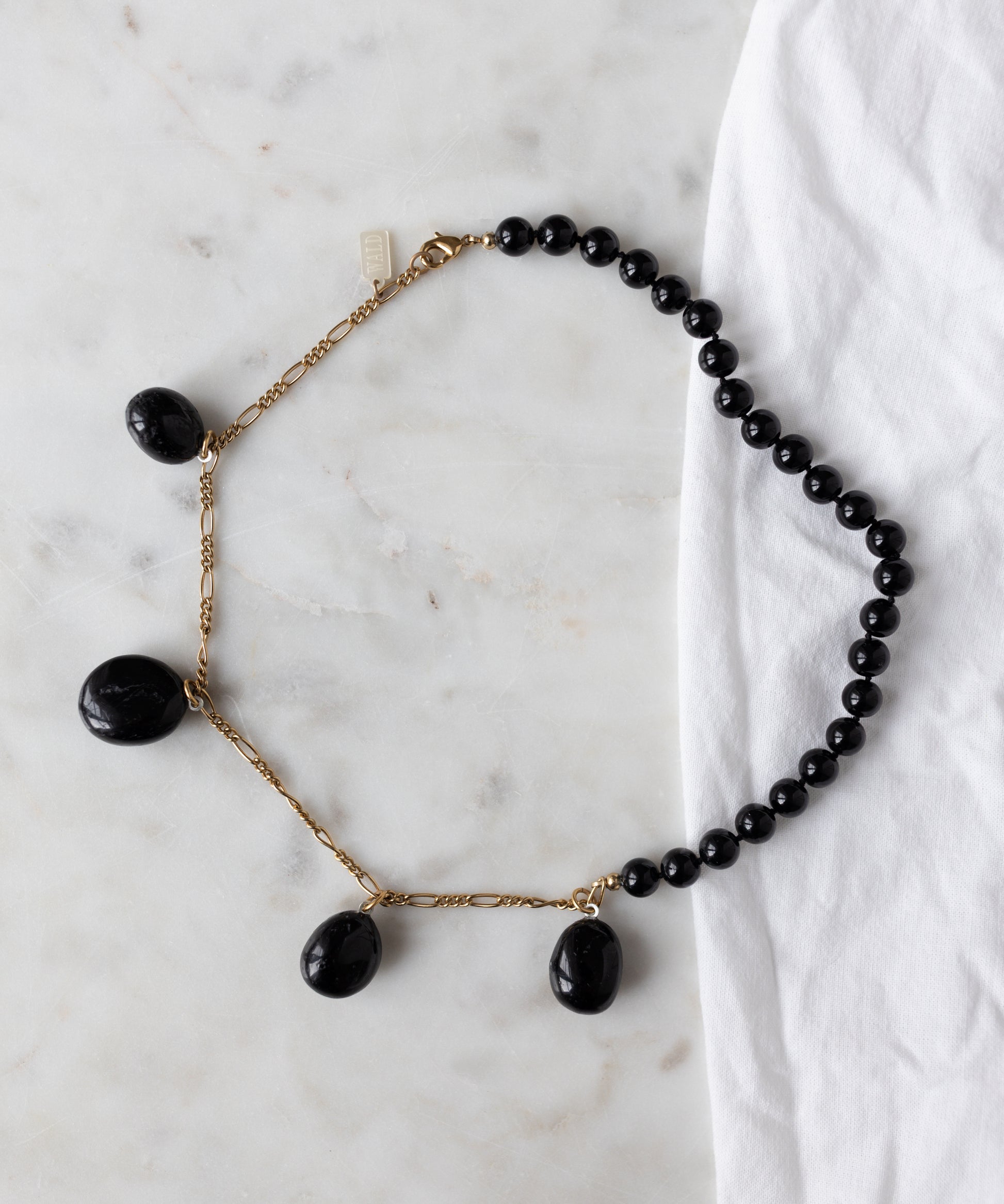 The WALD Berlin Collection Noir Pas De Deux Black Drop Necklace features three large black pendants and an 18k gold-plated chain, shown on white marble beside white fabric.