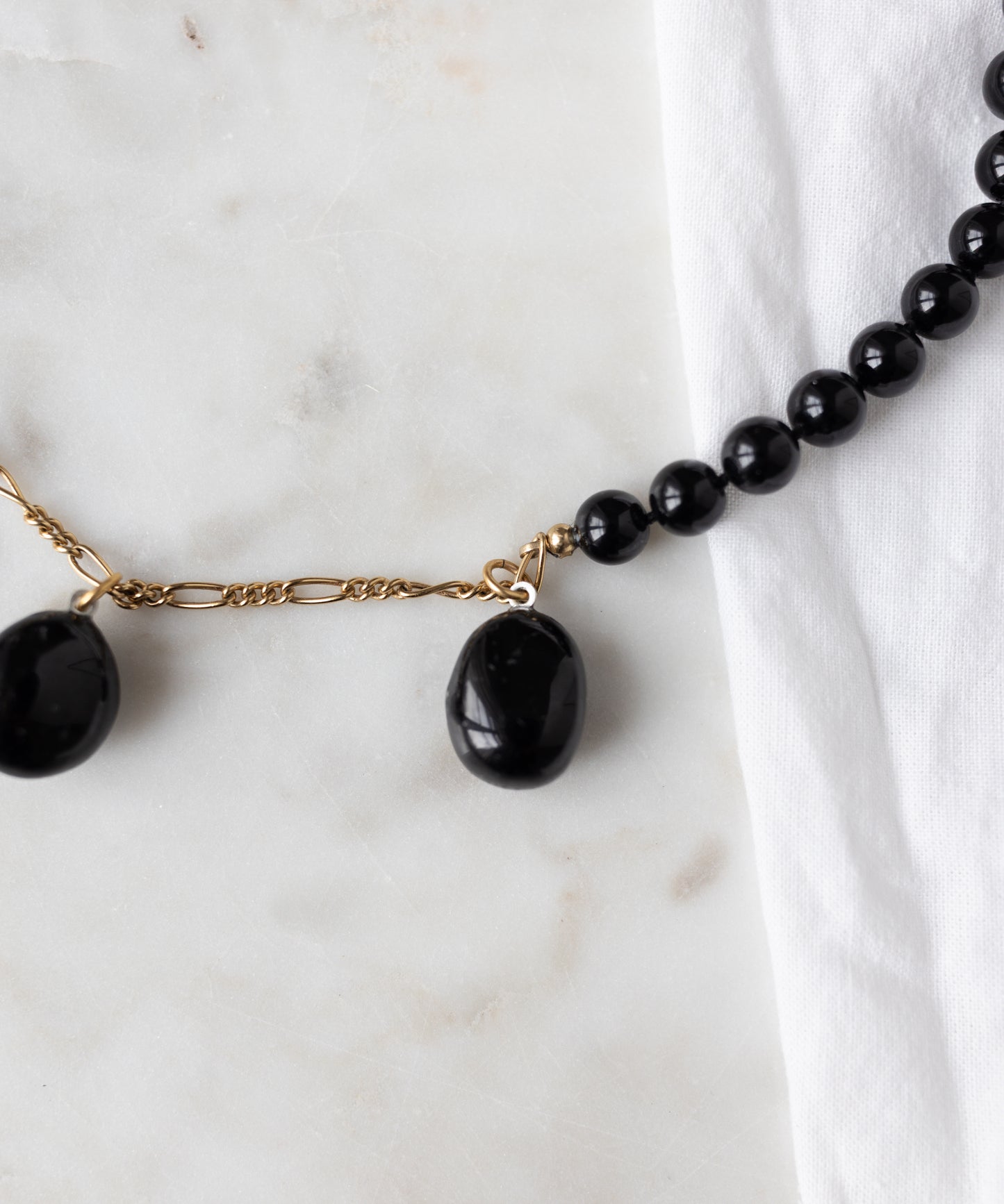 A close-up of WALD Berlin's Collection Noir Pas De Deux Black Drop Necklace, featuring an 18k gold plated clasp, is displayed on white marble beside a white cloth.