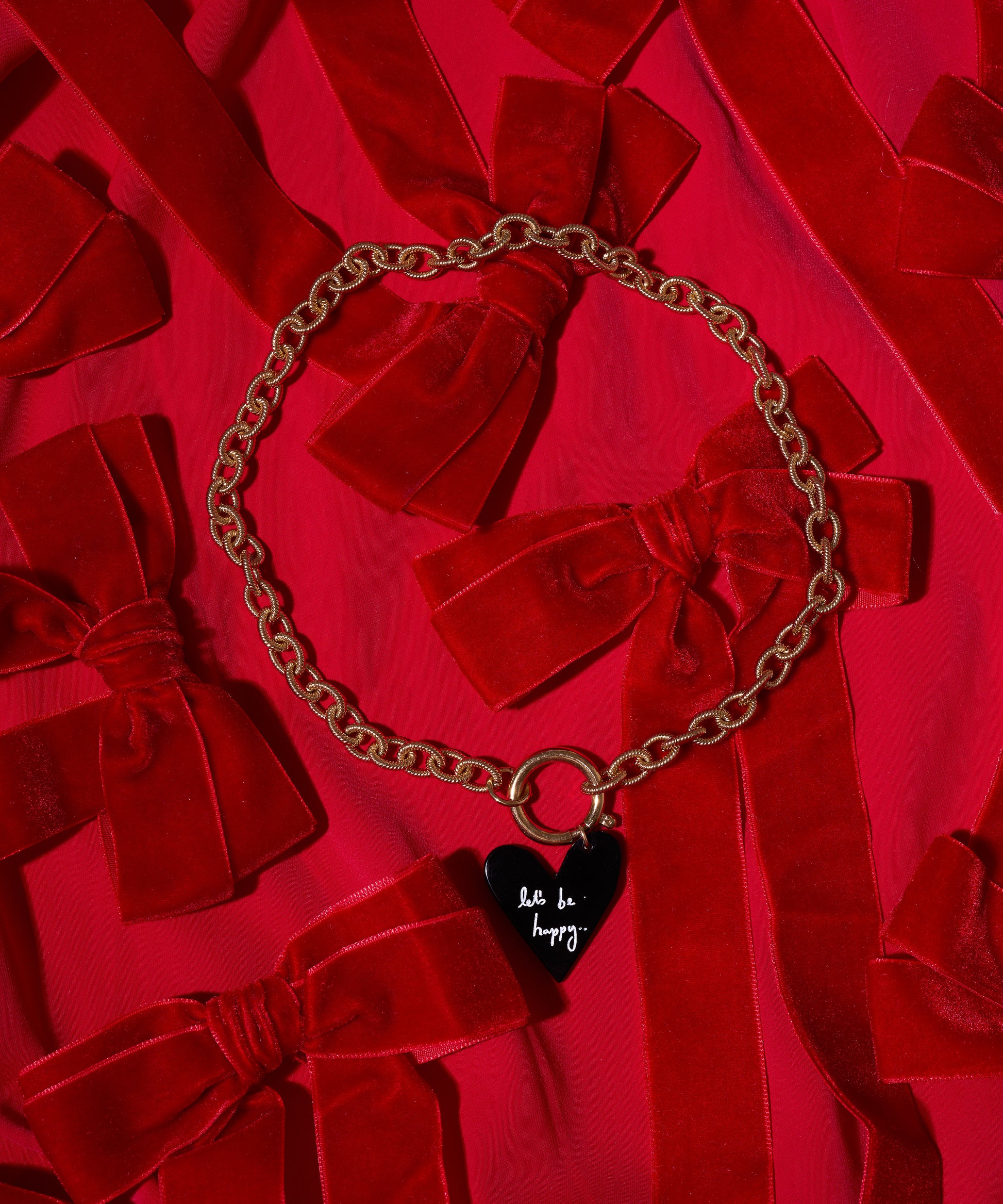 The WALD Berlin Collection Noir Let’s Be Happy Gold Necklace features an 18k gold-plated chain and a black heart pendant with “be happy,” displayed on red fabric with velvet bows.