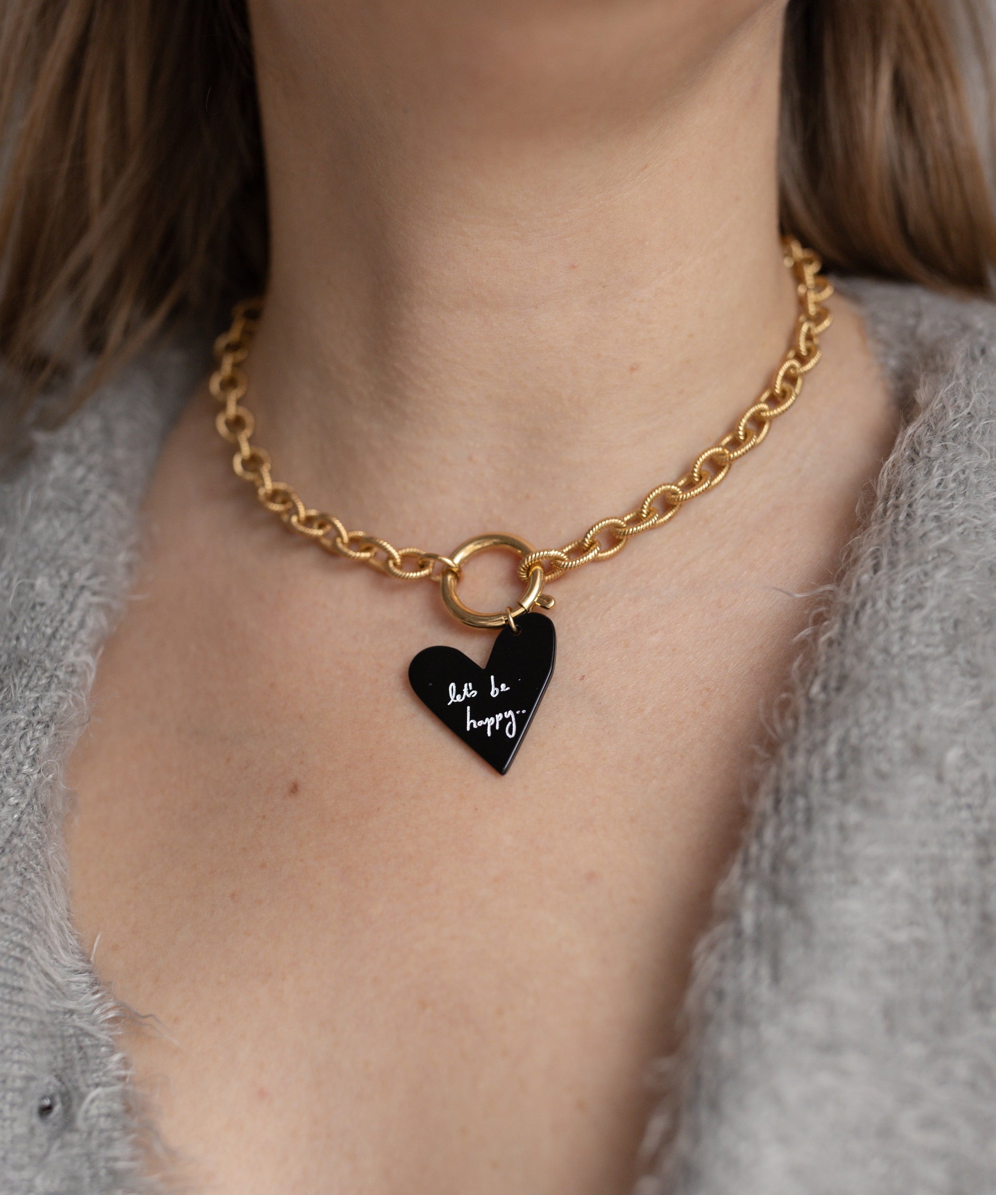 A person in a gray fuzzy sweater wears the WALD Berlin Collection Noir Let’s Be Happy Gold Necklace, 18k gold plated, featuring a black heart pendant inscribed with "be happy.