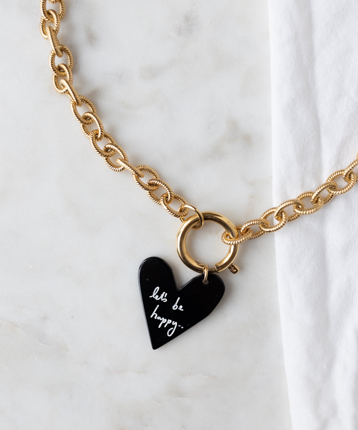 The WALD Berlin Collection Noir Let’S Be Happy Gold Necklace, 18k gold plated with a black heart "let's be happy..." pendant and round clasp, rests on light marble beside a white cloth.