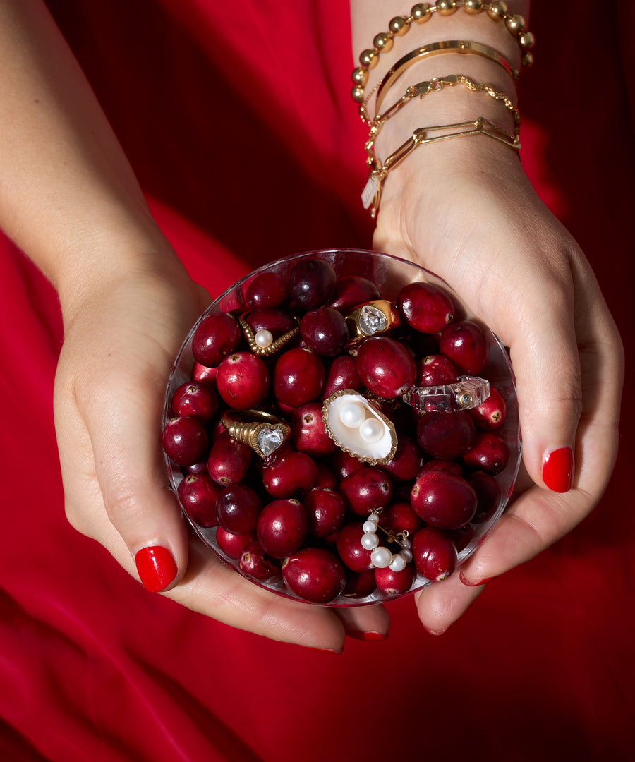 A person with red nail polish and gold bracelets holds a bowl of cranberries mixed with WALD Berlin's NYC If I Rule The World Ring and pearl jewelry against a red background.
