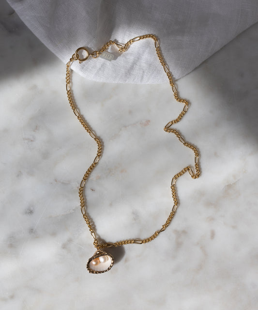 The Drop It Like It’s Hot Shell Necklace by WALD Berlin is a handmade gold-plated necklace with a delicate pearl pendant, displayed on marble with soft fabric and gentle shadows in the background.
