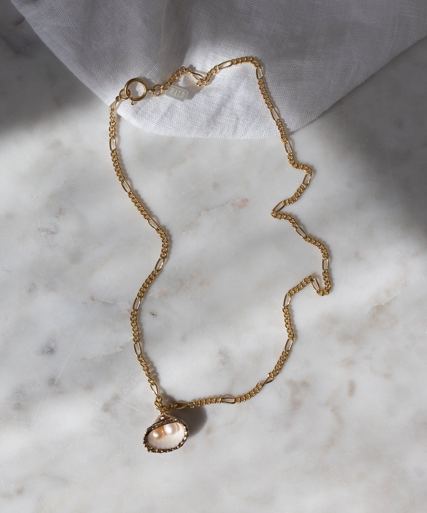 The Drop It Like It’s Hot Shell Necklace by WALD Berlin is a handmade gold-plated necklace with a delicate pearl pendant, displayed on marble with soft fabric and gentle shadows in the background.