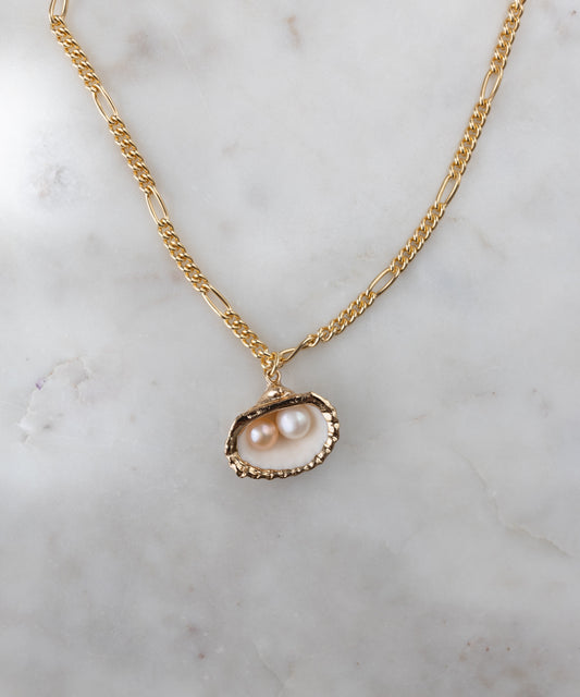 The Drop It Like It’s Hot Shell Necklace by WALD Berlin features a gold-plated pendant with two pearls in a textured oval frame, displayed on white marble—a stunning piece of handmade jewelry.