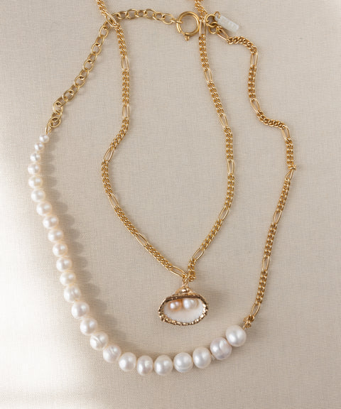 Two handmade WALD Berlin jewelry pieces are shown: the "Drop It Like It's Hot Shell Necklace," a gold-plated piece with a pearl shell pendant, and a pearl strand necklace, both set on a neutral fabric background.
