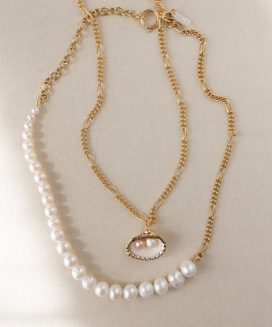 Two handmade WALD Berlin jewelry pieces are shown: the "Drop It Like It's Hot Shell Necklace," a gold-plated piece with a pearl shell pendant, and a pearl strand necklace, both set on a neutral fabric background.