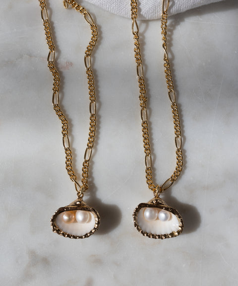 Two WALD Berlin Drop It Like It's Hot Shell Necklaces, gold-plated and handmade with shell pendants holding two small pearls each, are displayed on a light marble surface—a modern spin on the classic pearl necklace.