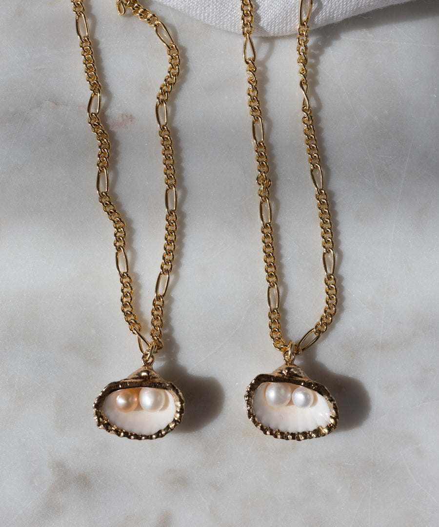 Two WALD Berlin Drop It Like It's Hot Shell Necklaces, gold-plated and handmade with shell pendants holding two small pearls each, are displayed on a light marble surface—a modern spin on the classic pearl necklace.
