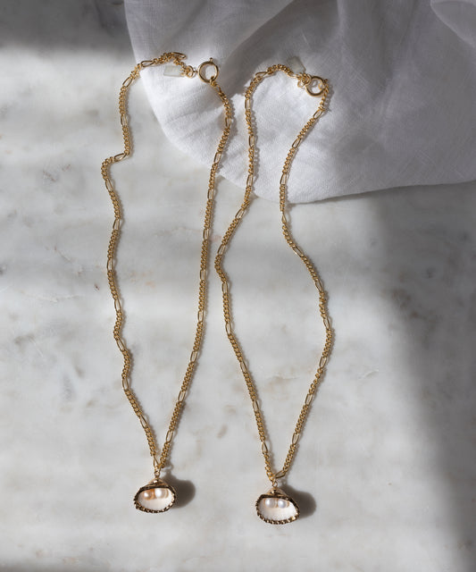 The Drop It Like It’s Hot Shell Necklace set by WALD Berlin features two handmade gold-plated chains with oval pearl pendants, elegantly displayed on marble near a white cloth, radiating timeless charm.