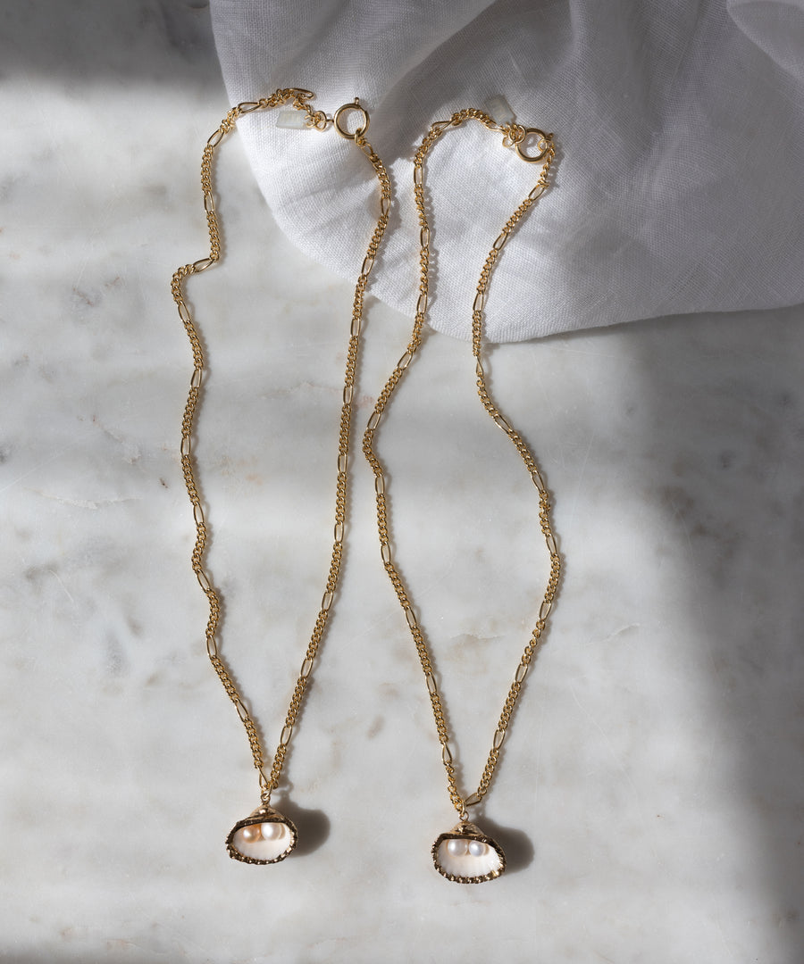 The Drop It Like It’s Hot Shell Necklace set by WALD Berlin features two handmade gold-plated chains with oval pearl pendants, elegantly displayed on marble near a white cloth, radiating timeless charm.