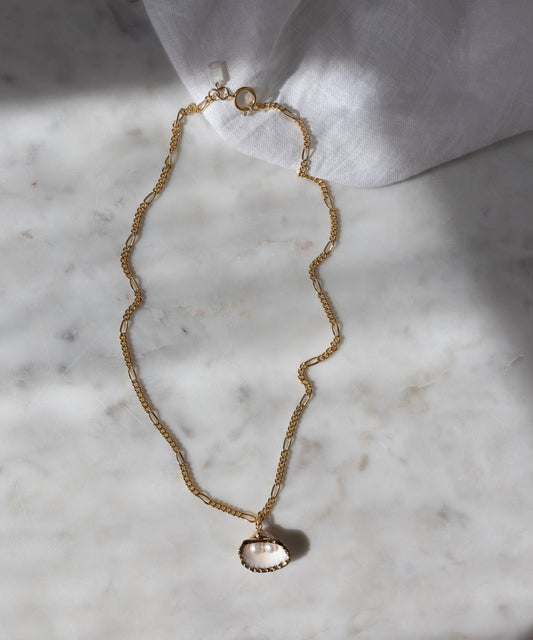 The WALD Berlin Drop It Like It’s Hot Shell Necklace, a gold-plated piece with a shell-shaped pendant, is displayed on marble with white fabric in the corner, highlighting this elegant handmade jewelry.