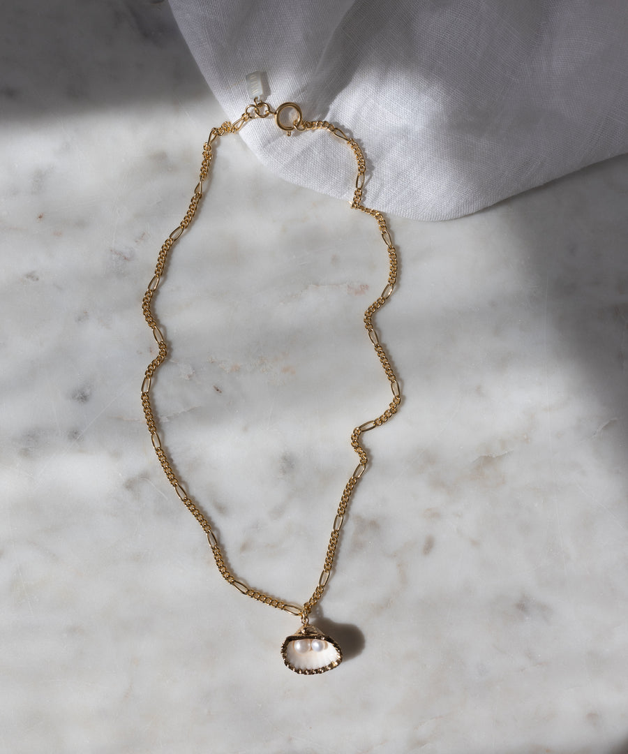 The WALD Berlin Drop It Like It’s Hot Shell Necklace, a gold-plated piece with a shell-shaped pendant, is displayed on marble with white fabric in the corner, highlighting this elegant handmade jewelry.