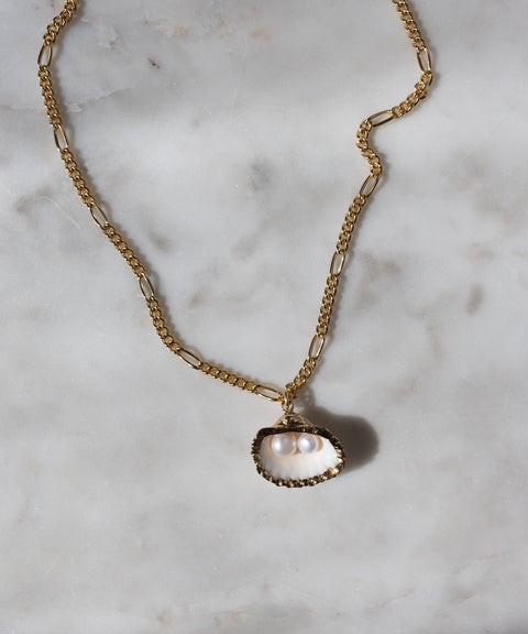 The WALD Berlin "Drop It Like It’s Hot Shell Necklace" is a gold-plated, handmade piece featuring an oyster shell pendant with two white pearls, elegantly displayed on white marble.