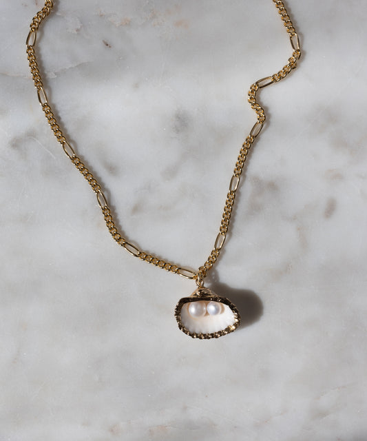 The WALD Berlin "Drop It Like It’s Hot Shell Necklace" is a gold-plated, handmade piece featuring an oyster shell pendant with two white pearls, elegantly displayed on white marble.