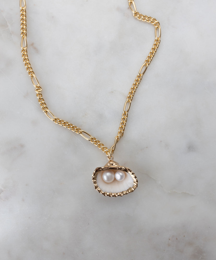 The Drop It Like It’s Hot Shell Necklace by WALD Berlin features an elegant gold-plated brass chain with an open oyster shell pendant and two pearls, shown handmade against a light marble background.