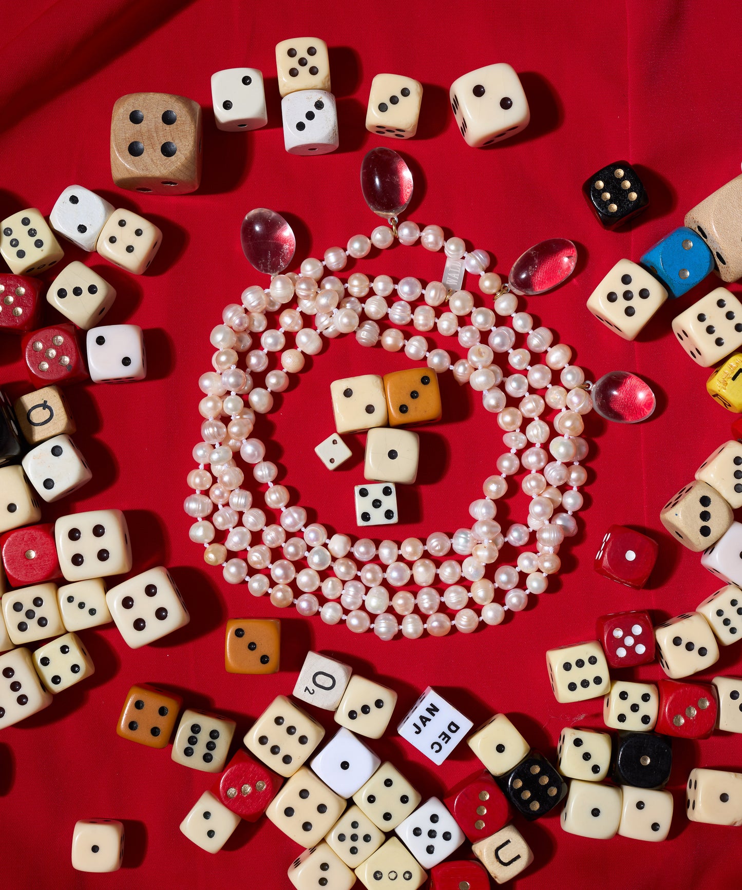 The WALD Berlin Happy Holidays Long Pearl Drop Necklace is displayed on red fabric, featuring sweetwater pearls, berg crystal pendants, assorted dice, and a few pink gems.