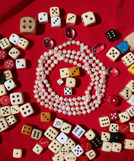 The WALD Berlin Happy Holidays Long Pearl Drop Necklace is displayed on red fabric, featuring sweetwater pearls, berg crystal pendants, assorted dice, and a few pink gems.