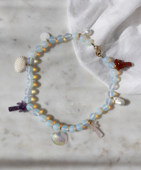 The Mexicana La Bamba Necklace by WALD Berlin features handmade iridescent opalite beads, shell, pearl, and stone cross charms—jewelry inspired by Mexicana style and displayed on marble with a white cloth.