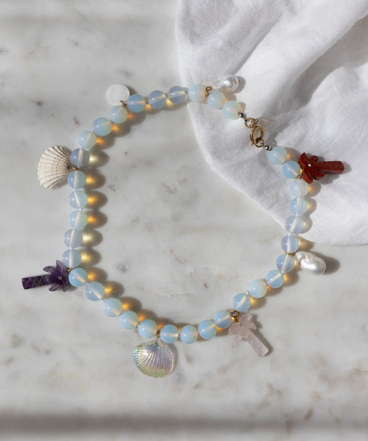 The Mexicana La Bamba Necklace by WALD Berlin features handmade iridescent opalite beads, shell, pearl, and stone cross charms—jewelry inspired by Mexicana style and displayed on marble with a white cloth.