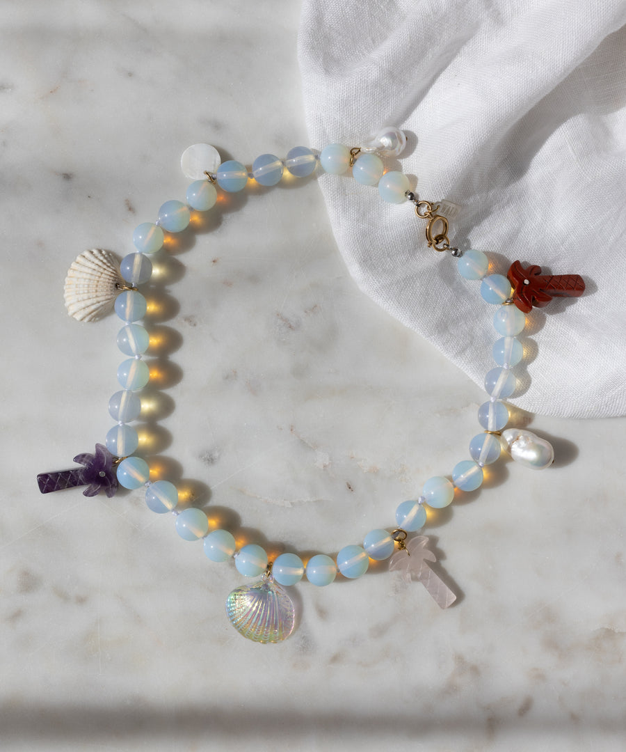 The Mexicana La Bamba Necklace by WALD Berlin features handmade iridescent opalite beads, shell, pearl, and stone cross charms—jewelry inspired by Mexicana style and displayed on marble with a white cloth.