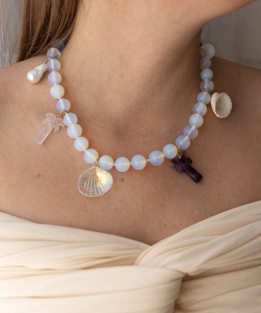 Wearing the WALD Berlin Mexicana La Bamba Necklace, which features opalite beads and palm tree, cross, shell, and seashell pendants, paired with an off-shoulder cream top.