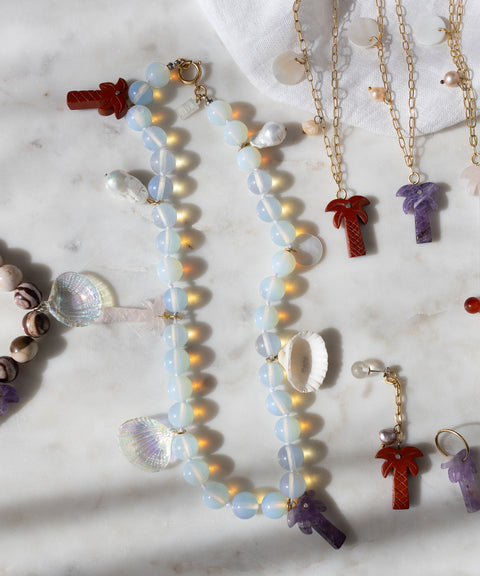Handmade jewelry by WALD Berlin, like the Mexicana La Bamba Necklace with opalite beads, plus gemstone, shell, and palm tree charm bracelets and earrings—all beautifully displayed on a marble surface.