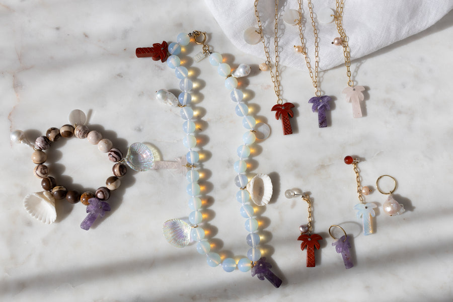 WALD Berlin Mexicana Mariachi Earrings Pearl, displayed with beaded necklaces, bracelets, baroque pearl earrings, and gold chain necklaces with palm tree pendants on marble among shells and pearls. Some pieces are handmade in Germany.