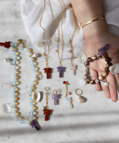 A hand with gold bracelets displays WALD Berlin handmade jewelry, including the Mexicana La Bamba Necklace with opalite beads, plus bracelets, earrings, and keychains featuring cross, shell, and palm tree pendants on a marble surface.