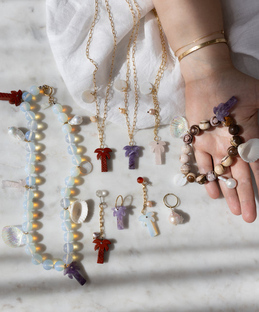 A hand with gold bracelets displays WALD Berlin handmade jewelry, including the Mexicana La Bamba Necklace with opalite beads, plus bracelets, earrings, and keychains featuring cross, shell, and palm tree pendants on a marble surface.