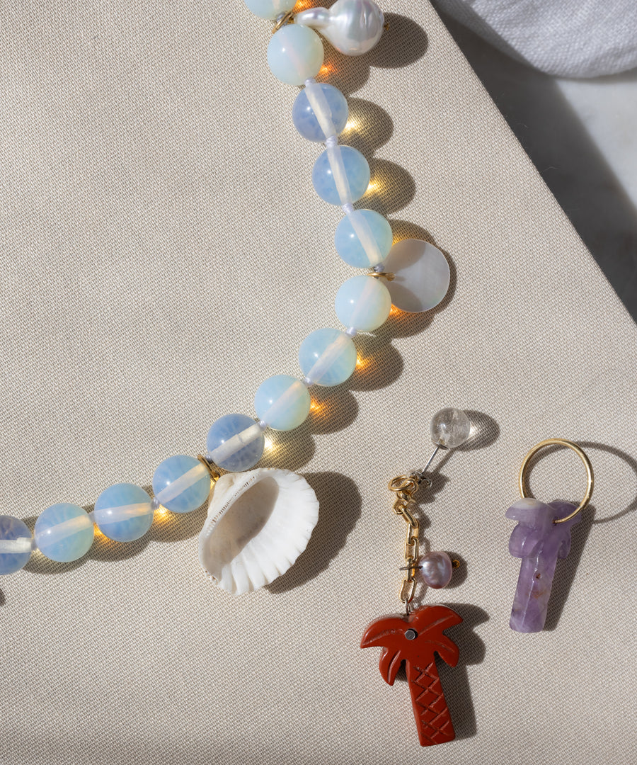 The WALD Berlin Mexicana La Bamba Necklace, a red palm tree keychain, and a purple crystal charm keyring rest on beige fabric—a perfect trio for handmade jewelry lovers.