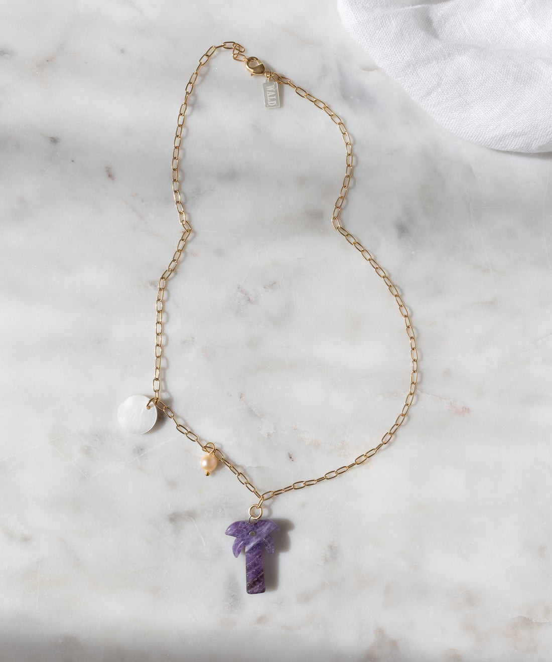 Die WALD Berlin Mexicana Solo Amore Halskette ist ein handgefertigtes, vergoldetes Stück mit einem violetten Steinanhänger, einem runden weißen Charm und einer kleinen Perle, die elegant auf weißem Marmor präsentiert werden.