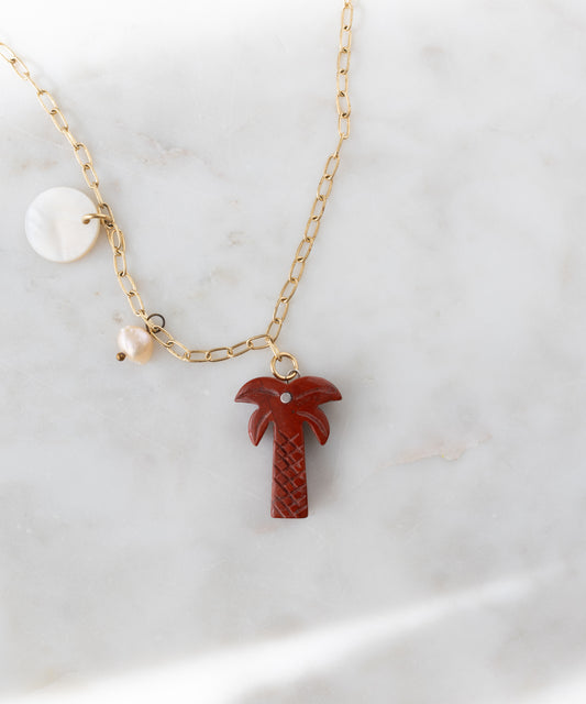 Die WALD Berlin Mexicana Solo Amore Halskette ist ein handgefertigtes, vergoldetes Stück mit einem handgeschnitzten Palmenanhänger aus Stein, einem runden weißen Anhänger und einem Perlenanhänger, der elegant auf weißem Marmor präsentiert wird.