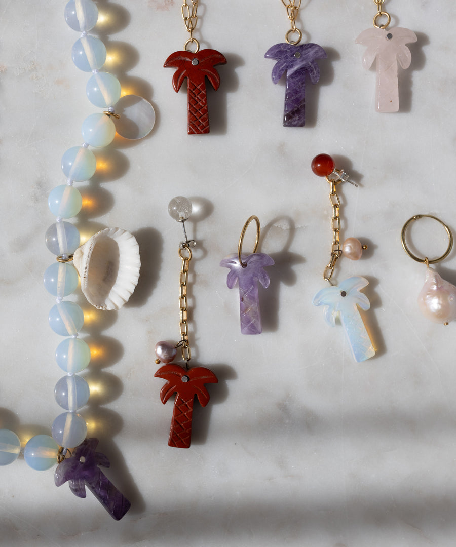 WALD Berlin’s Mexicana Mariachi Earrings Pearl and other palm tree necklaces, colorful earrings, a pearl ring, and a seashell are displayed on marble with handmade asymmetric gold earrings from Germany.