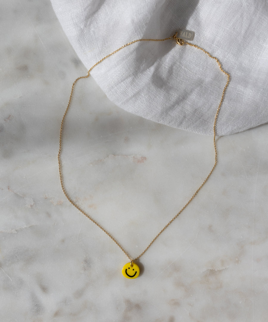WALD Berlin’s Smilie Yellow Necklace, a gold-plated piece featuring a hand-painted pendant, is elegantly displayed on a marble surface with a white cloth in the background.