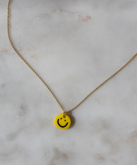 WALD Berlin’s Smilie Yellow Necklace features a gold-plated, hand-painted pendant with a cheerful yellow smiley face, elegantly showcased on a light marble surface.