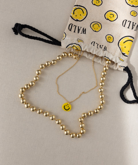 A gold-plated necklace and a gold chain with a yellow smiley face pendant from WALD Berlin are displayed near a drawstring bag adorned with yellow smiley faces and the word “WILD.” This playful arrangement is highlighted by the Smilie Yellow Necklace from WALD Berlin, featuring a charming, hand-painted pendant.