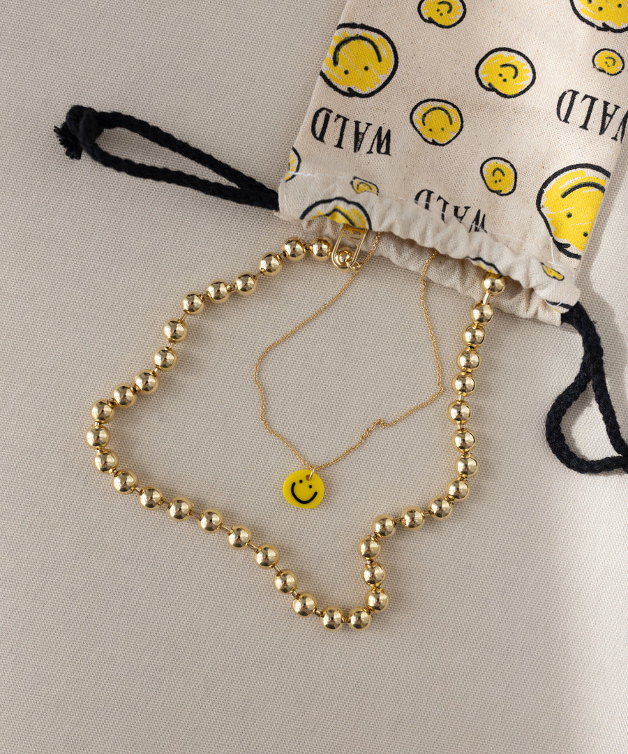 A gold-plated necklace and a gold chain with a yellow smiley face pendant from WALD Berlin are displayed near a drawstring bag adorned with yellow smiley faces and the word “WILD.” This playful arrangement is highlighted by the Smilie Yellow Necklace from WALD Berlin, featuring a charming, hand-painted pendant.