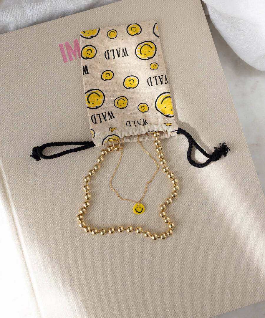 A beige surface displays a drawstring pouch adorned with smiley faces, a gold beaded necklace, and the WALD Berlin Smilie Yellow Necklace—a gold-plated accessory featuring a hand-painted pendant.
