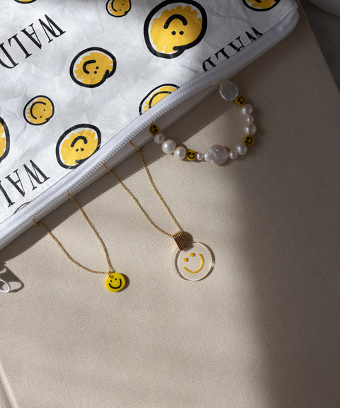 A WALD Berlin Smilie Yellow Necklace is displayed on a beige surface alongside a white pouch decorated with yellow smiley faces, a gold-plated necklace featuring a smiley pendant, a clear hand-painted pendant necklace, and a pearl bracelet adorned with small smiley beads.
