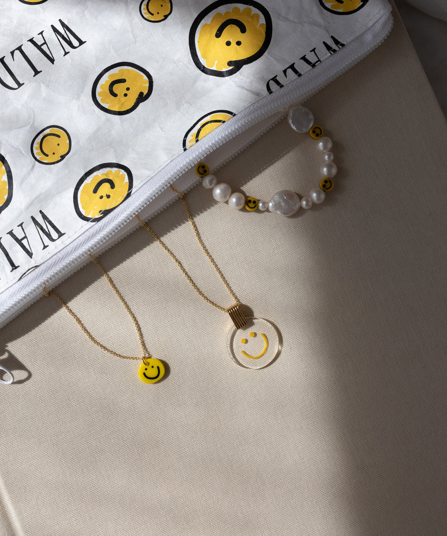A WALD Berlin Smilie Yellow Necklace is displayed on a beige surface alongside a white pouch decorated with yellow smiley faces, a gold-plated necklace featuring a smiley pendant, a clear hand-painted pendant necklace, and a pearl bracelet adorned with small smiley beads.