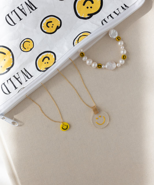 The WALD Berlin Smilie Yellow Necklace is arranged alongside a beaded bracelet and a clear hand-painted pendant with smiley faces, next to a white zipper pouch featuring a matching smiley design, all displayed on a beige surface.