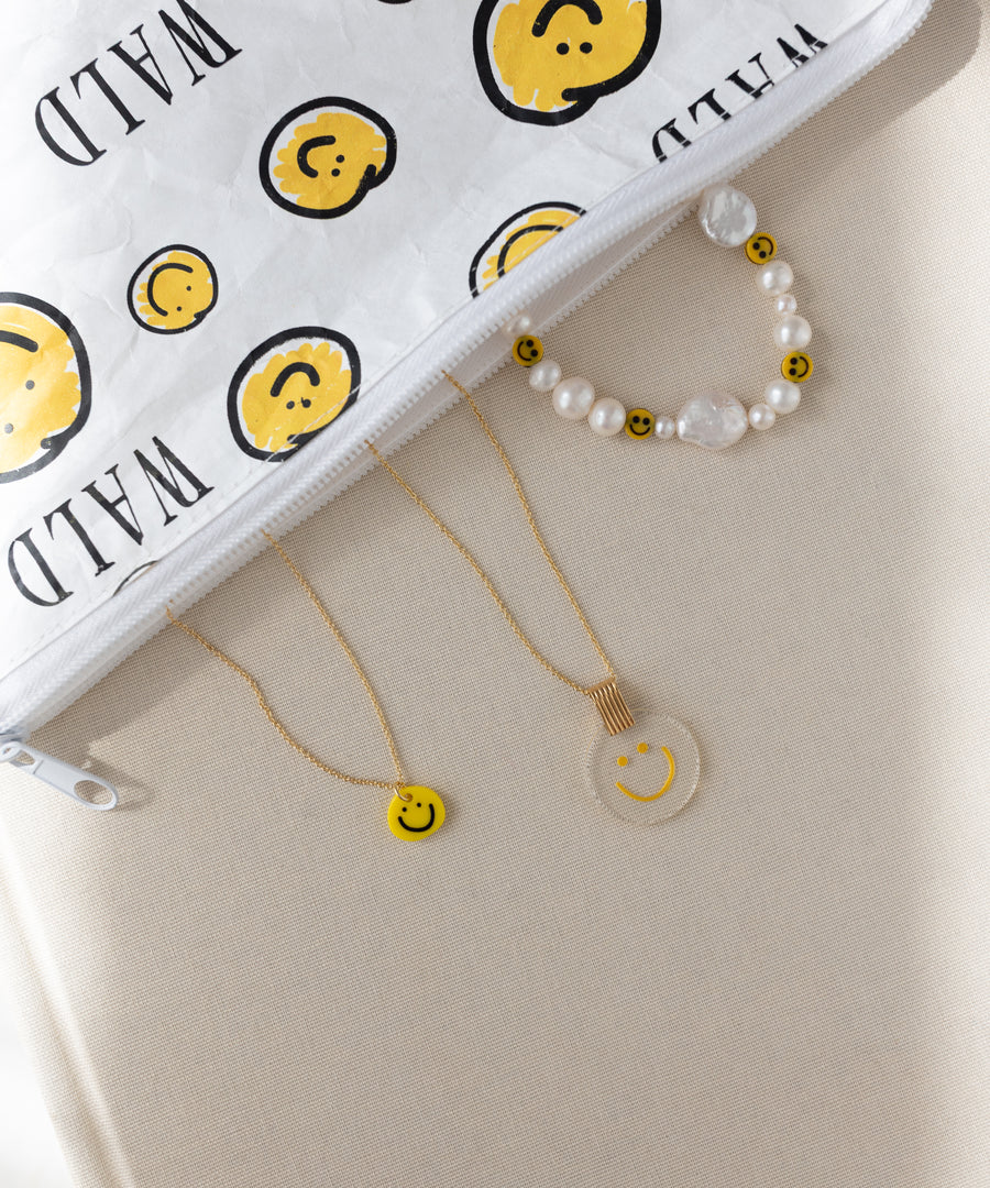 The WALD Berlin Smilie Yellow Necklace is arranged alongside a beaded bracelet and a clear hand-painted pendant with smiley faces, next to a white zipper pouch featuring a matching smiley design, all displayed on a beige surface.