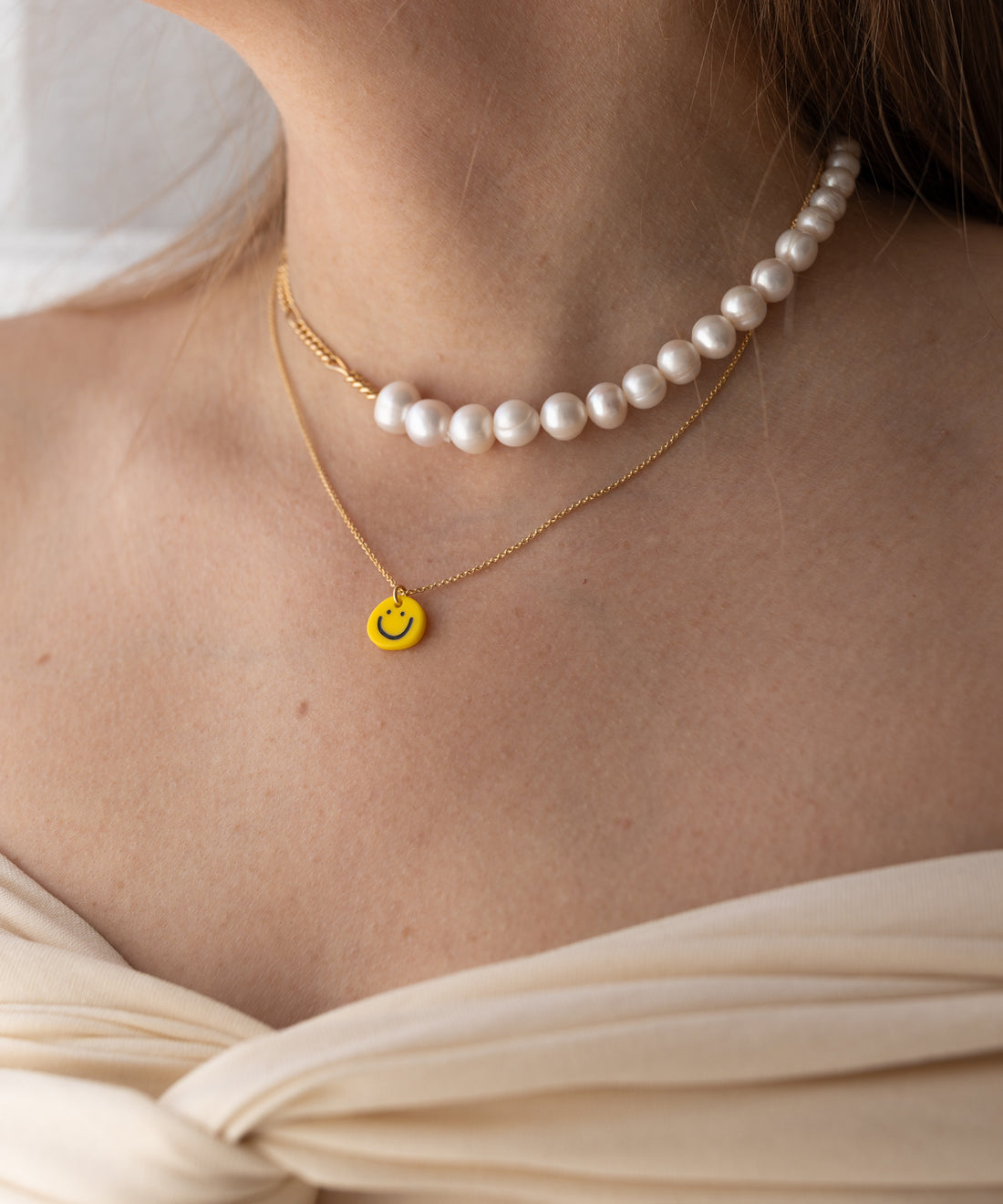 A person wearing a pearl necklace and the WALD Berlin Smilie Yellow Necklace—a gold-plated piece with a yellow smiley face pendant—paired with a cream-colored off-shoulder top.